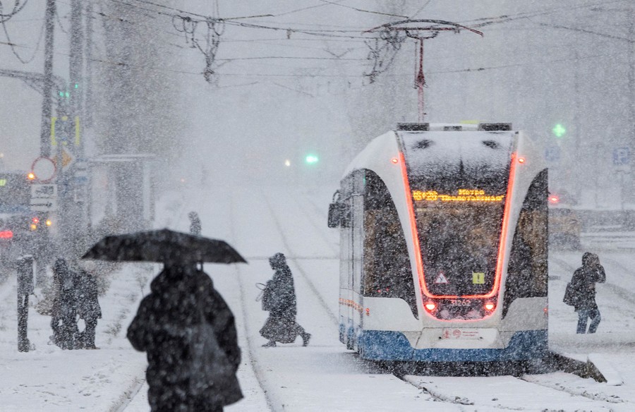 People cross a street behind a city tram on a snowy day.