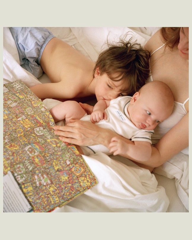 A toddler and baby lay in their mother's lap while she reads a book