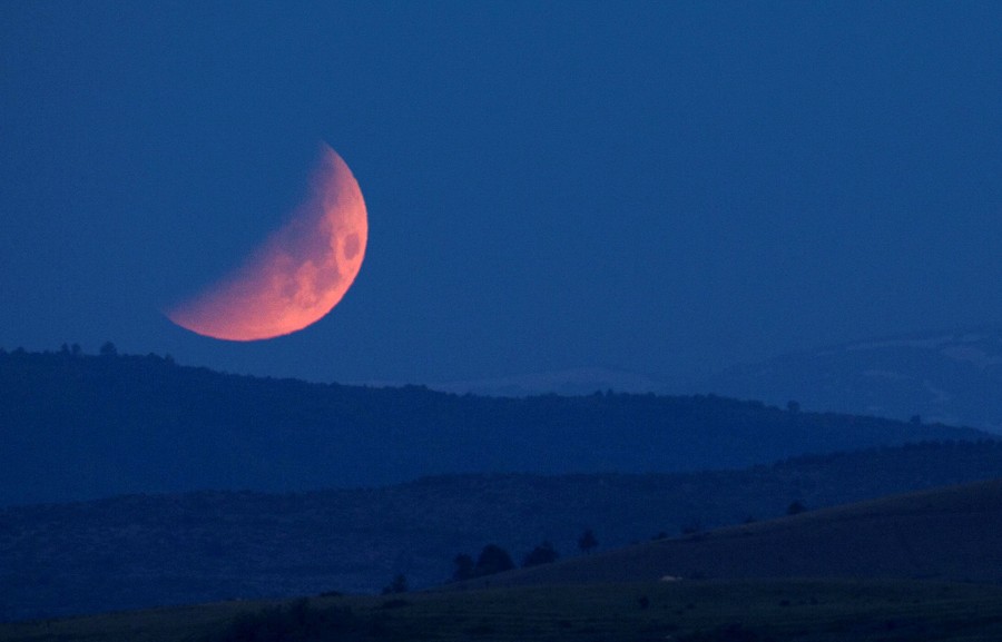 A view of a reddish-colored moon, half-obscured by the Earth's shadow, above distant hills.