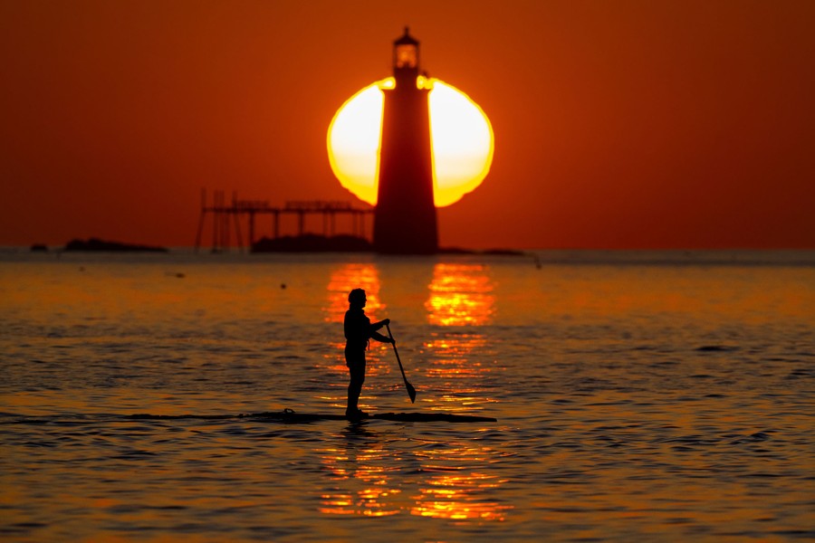 A person steers a stand-up paddleboard, with a lighthouse and sunrise in the background.