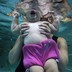 A baby is seen underwater from the chin down, held in the hands of an adult.