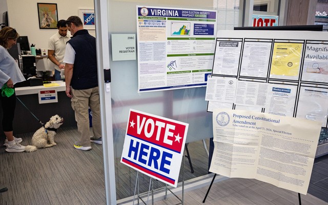 Photograph of an office with a red, white, and blue “VOTE HERE” sign with signs about election security, voting instructions, and a proposed constitutional amendment behind it