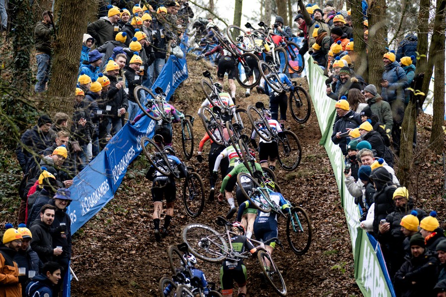 Spectators stand on either side of a steep race course, watching cyclists carrying their bikes up a hill.