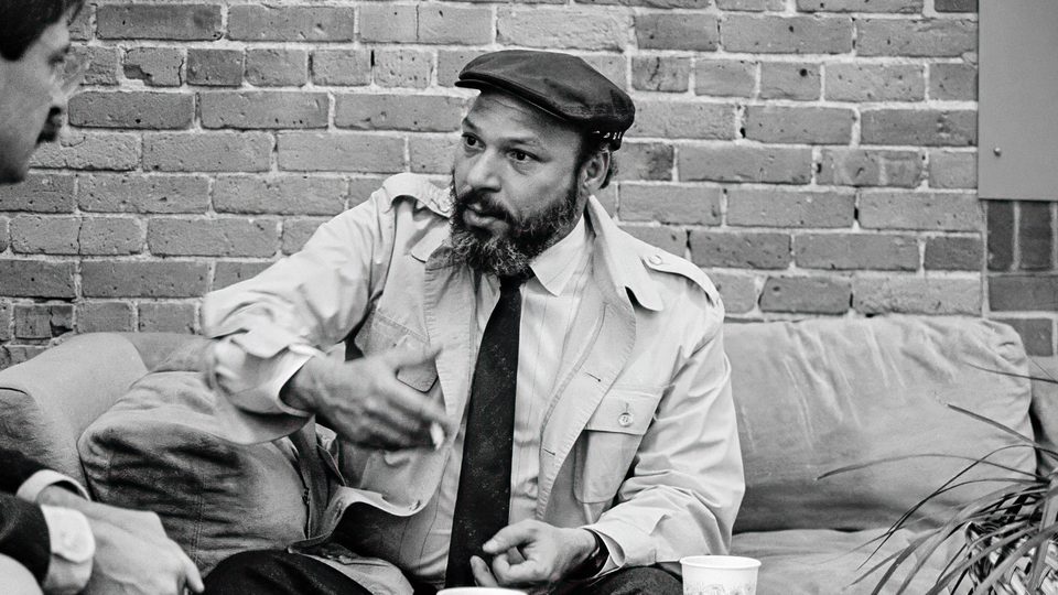 A black-and-white photo August Wilson in a trench coat, driving cap, and tie sitting on sofa and gesturing in conversation with a brick wall behind
