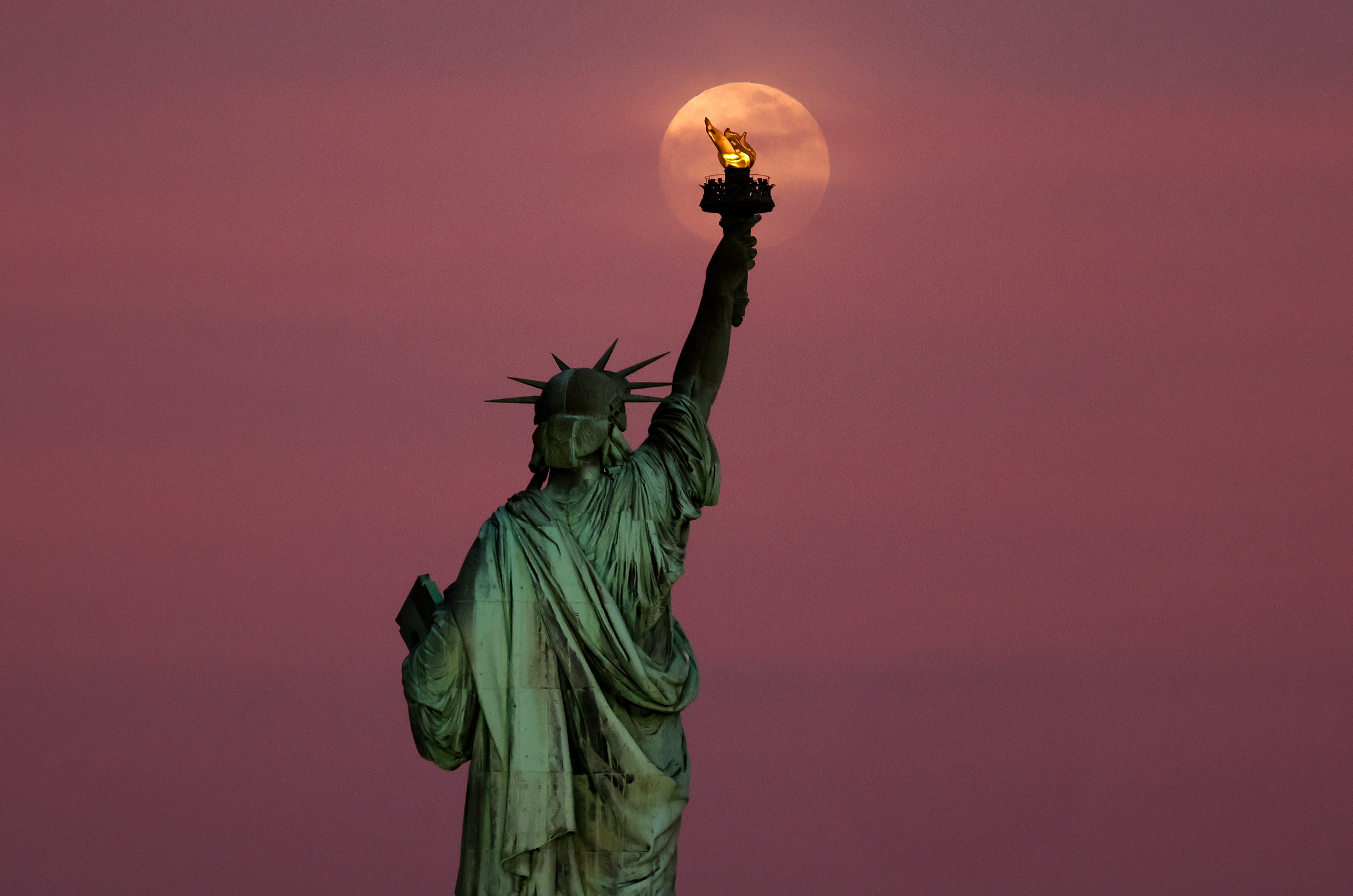 The moon rises, seen through clouds behind the Statue of Liberty.