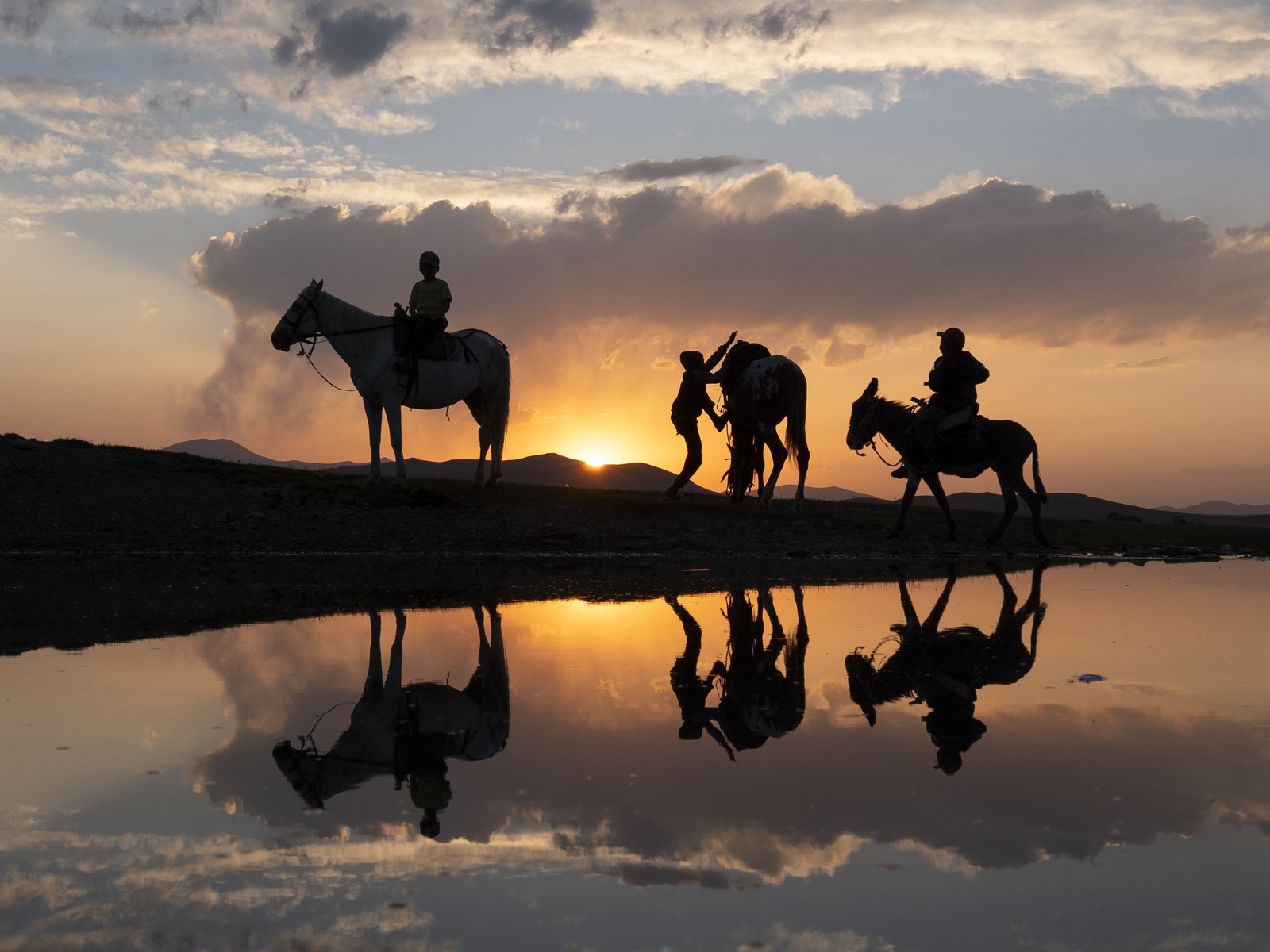 Three people ride on horses and donkeys, seen in silhouette and reflected in a pond at sunset.