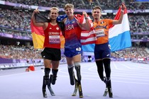 Three para-athletes wearing leg prostheses stand side by side, celebrating, draped in their countries' flags, inside a stadium.