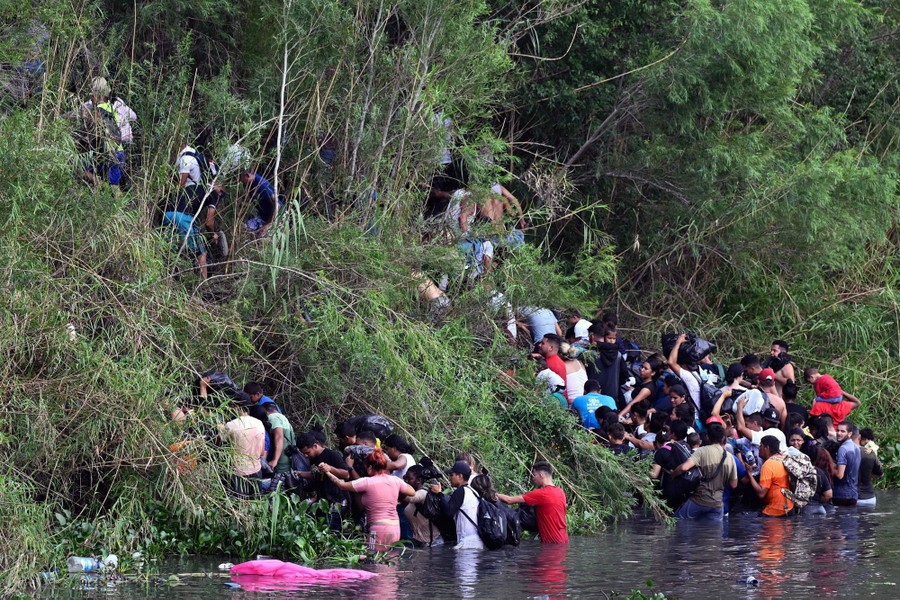 Dozens of people climb a steep hill after wading across a river.