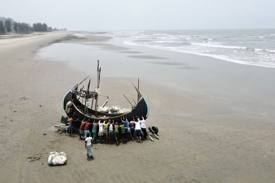 About 16 people push on the side of a small fishing boat on a wide beach.