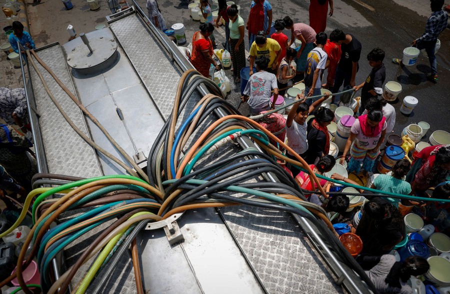 People fill containers with drinking water, using multiple hoses stuffed into an opening at the top of a tanker truck.