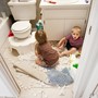 Two young children sit on the floor of a bathroom surrounded by packing peanuts. One child looks clearly alarmed and perhaps guilty of misbehavior.