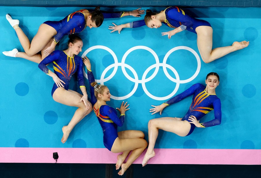 An overhead photo of five gymnasts lying down and posing around a set of Olympic rings printed on the floor
