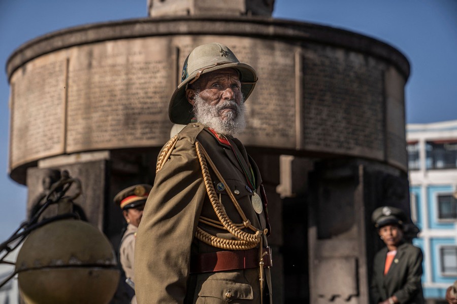 An older man stands beside a monument wearing a military uniform.