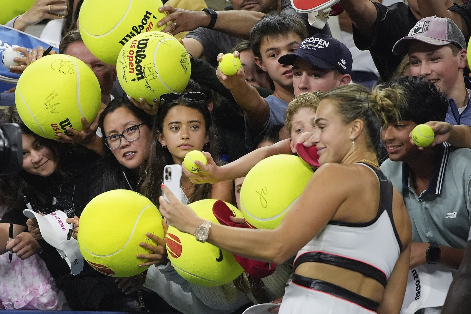 A tennis player takes selfies with fans in the stands, many of whom are holding large tennis balls.