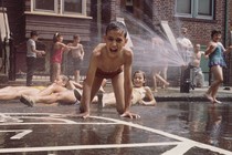 Children play in a city in front of rowhouses, in their swimwear, as a fire hydrant sprays water. One boy, front and center, seems excited and grins directly at the camera.