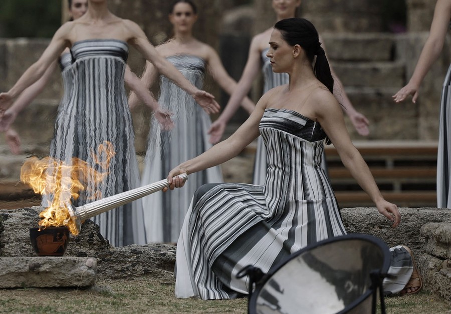 Several performers in long dresses pose while one kneels to light a torch.