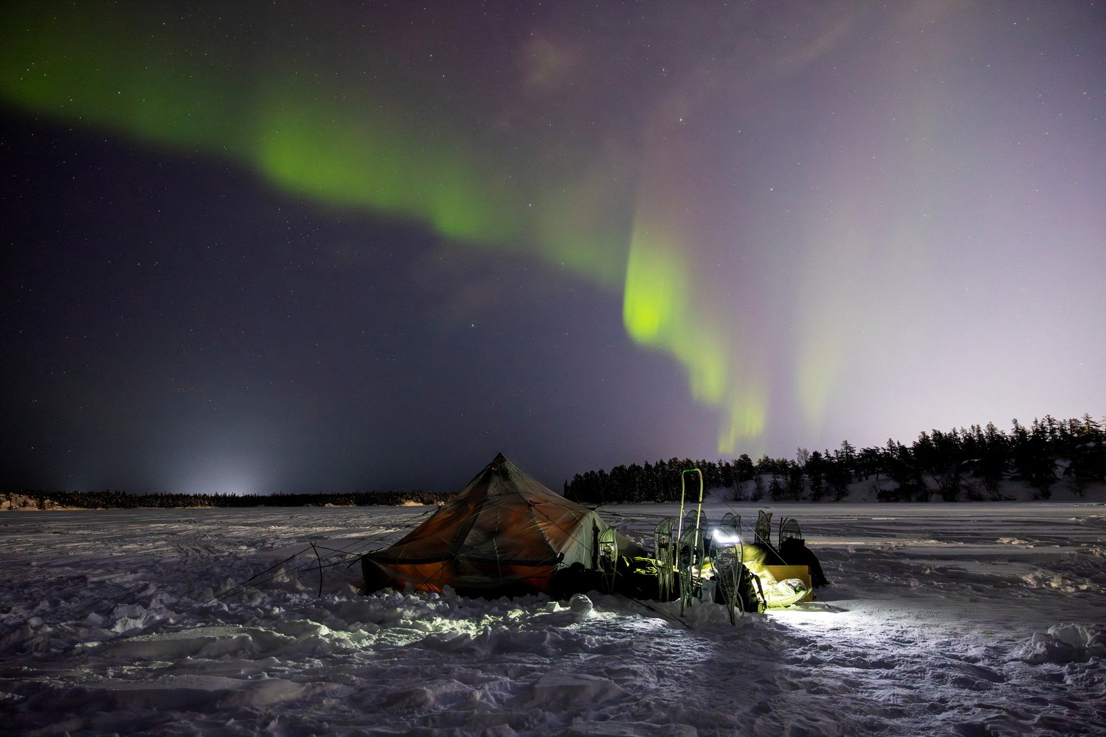 A tent sits beneath the Northern Lights, on an icy snow-covered plain.
