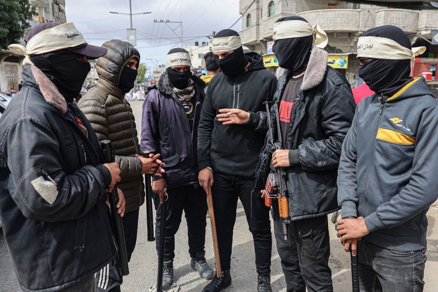 A group of six men stand in a street talking. Each wears a black balaclava over their face. One carries a rifle while the others carry long sticks.