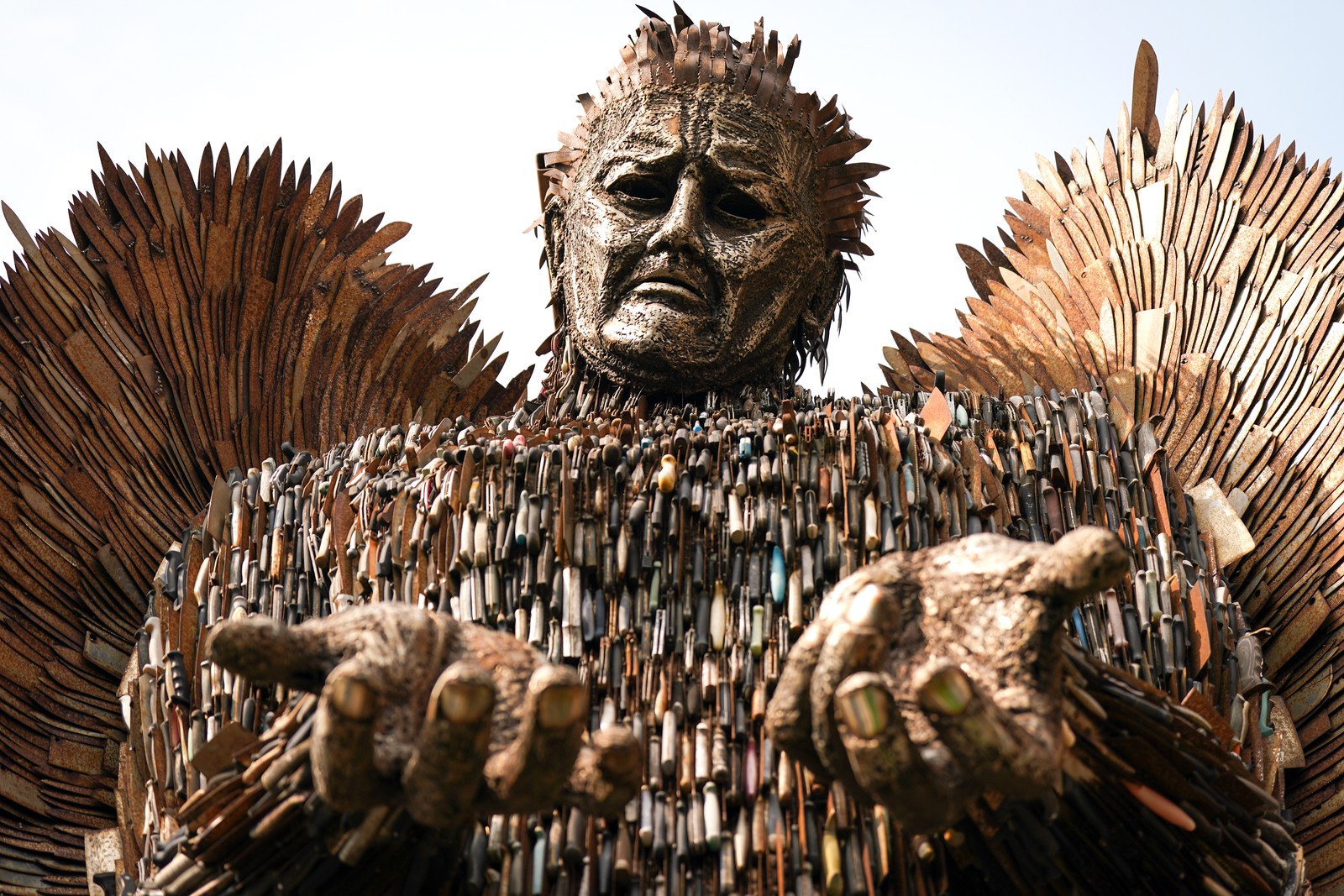 A large statue of an angel, made from the metal and blades of old knives, holding its upturned hands out.
