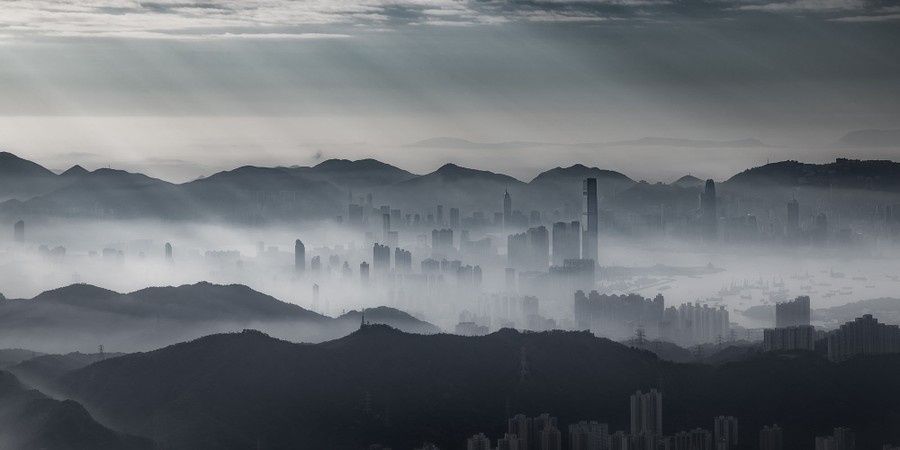 The buildings of Hong Kong stand amid mountains and mist in a wide landscape image.