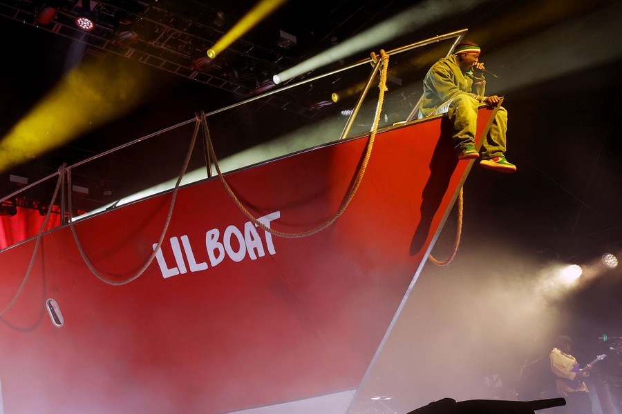 A performer sits on the bow of a life-size prop boat, with the name "LIL BOAT" written on the side.