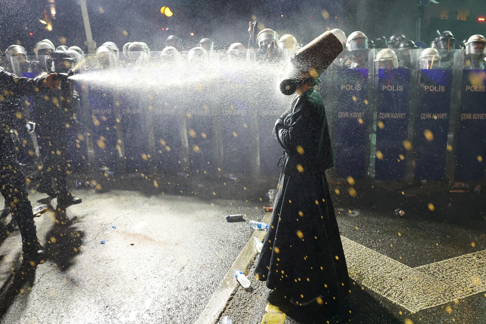 A protester wearing a traditional outfit and gas mask stands in front of a line of riot police, being sprayed in the face with pepper spray.