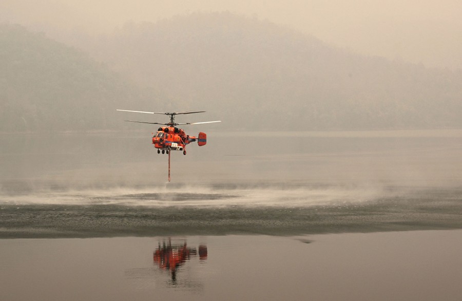 A firefighting helicopter hovers above a reservoir to suck water into a long hose.