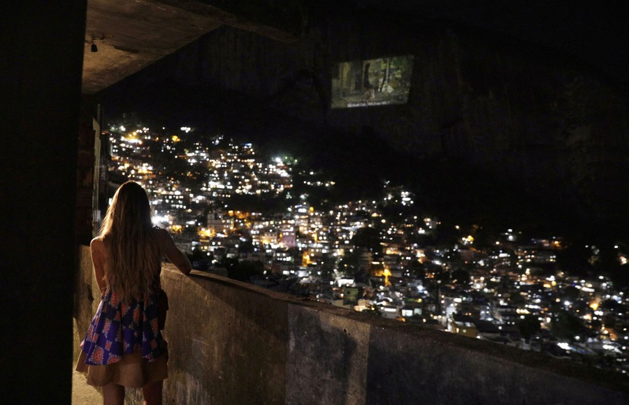A woman looks toward a movie projected on a nearby steep mountain face at night, above a lighted neighborhood.