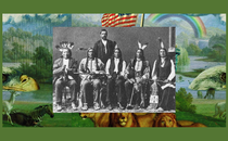 A black-and-white posed photograph of seated members of the Red Cloud delegation adorned in traditional regalia. Behind them stands a man in a suit.