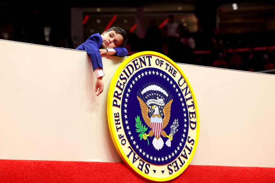 A child wearing a suit leans on a ledge above a large frame displaying the seal of the president of the United States.