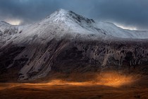 A distant view of a mountain with a dusting of early-season snow across the top, and a bright patch of sunlight on the valley floor below