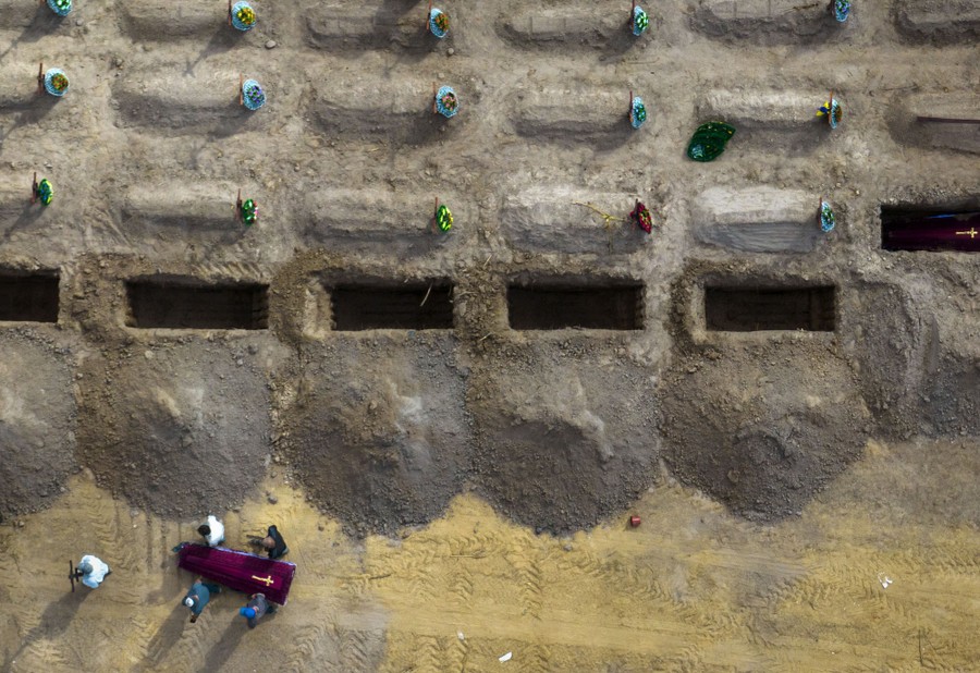 An aerial view of a cemetery with several open graves and people carrying a single coffin