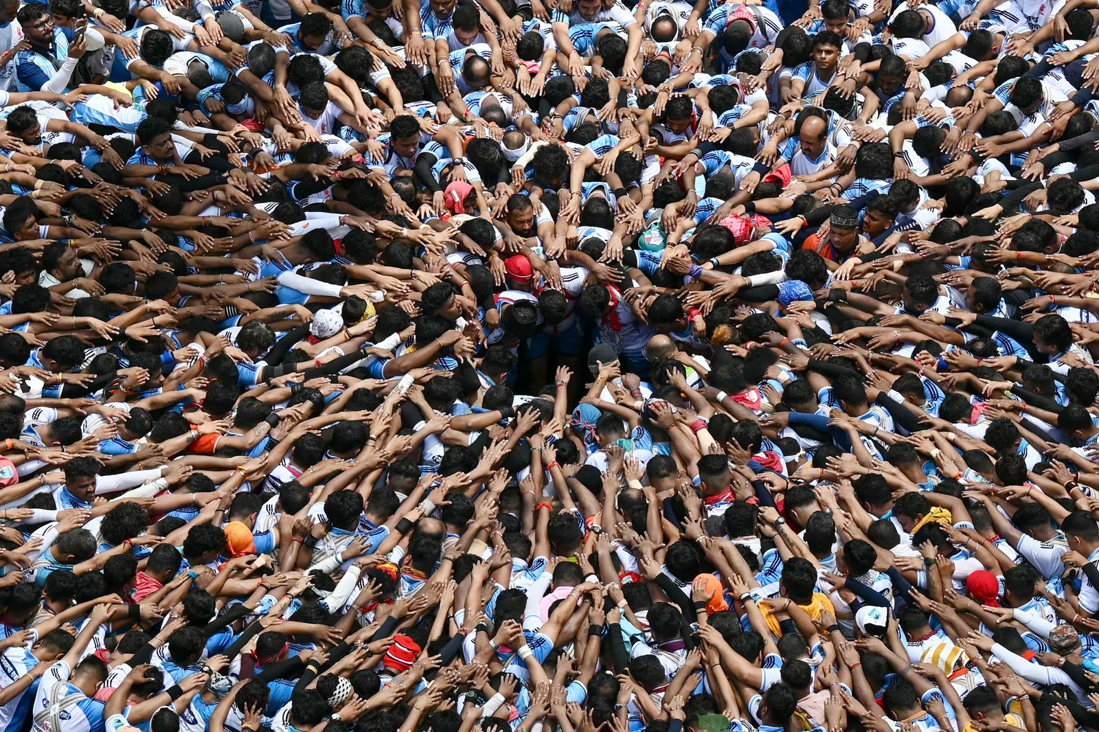A large crowd, seen from above, all reaching arms inward toward a small gap at center, where they will try to build a human pyramid.