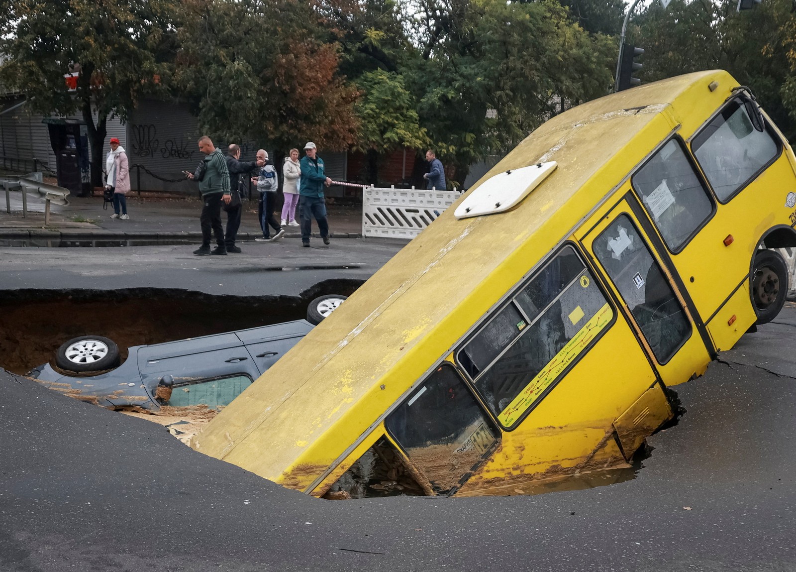 People walk past a sinkhole in a road with a bus and car in the hole.