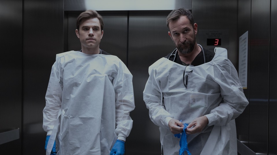 A photo of Patrick Ball and Noah Wyle standing in the elevator with medical gloves on