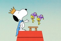 Atop his doghouse, Snoopy wears a crown and a cape as two small yellow birds in jester costumes hover.