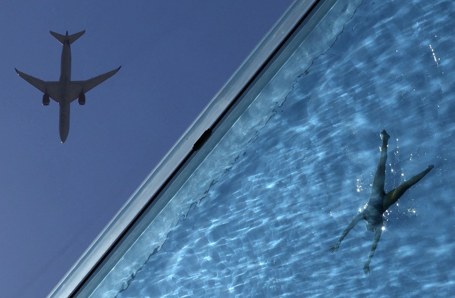 A view upward, partly through a transparent swimming pool, with an aircraft in the sky above.