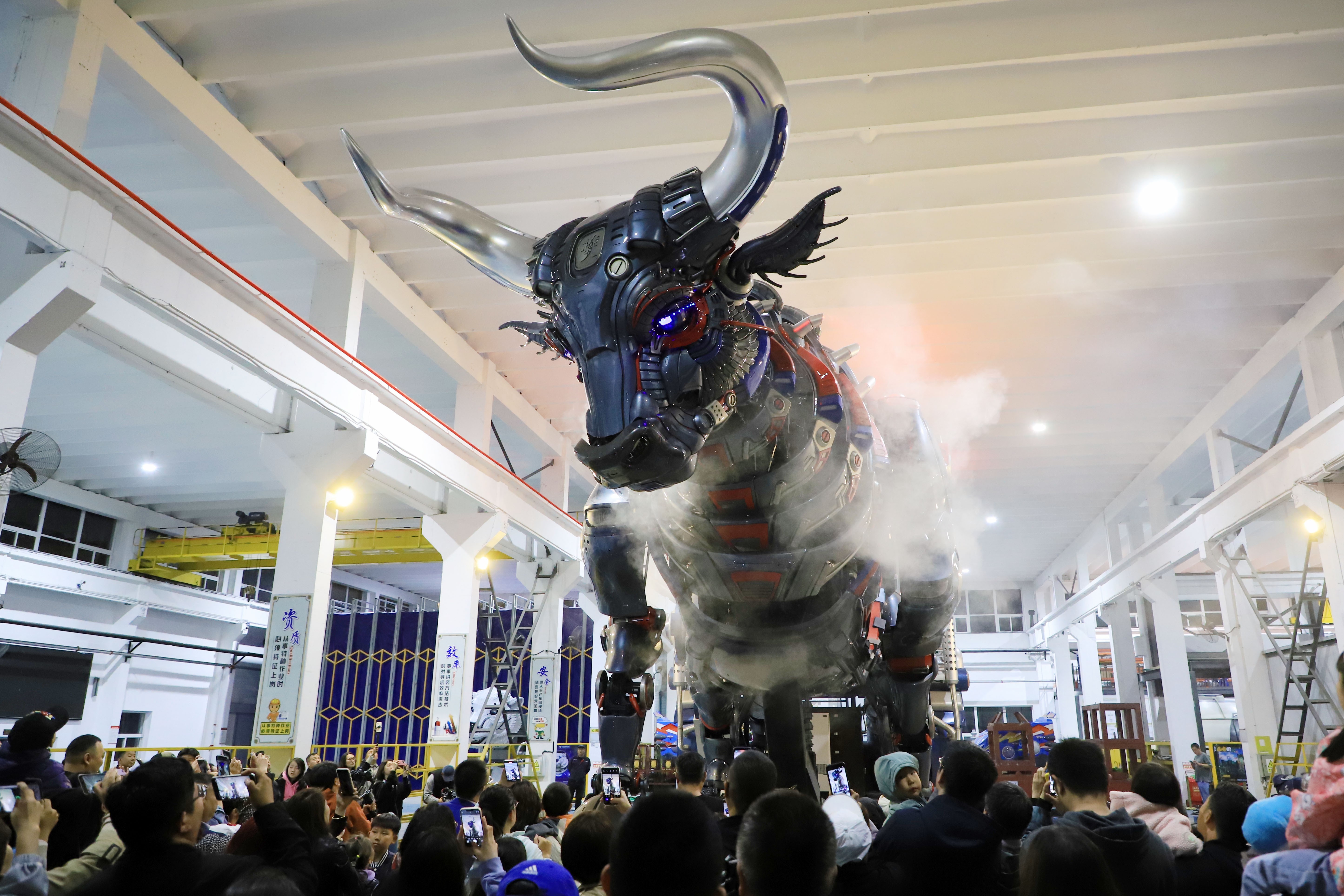 A crowd of people looks up at a large, mechanical puppet shaped like a mythical bull.