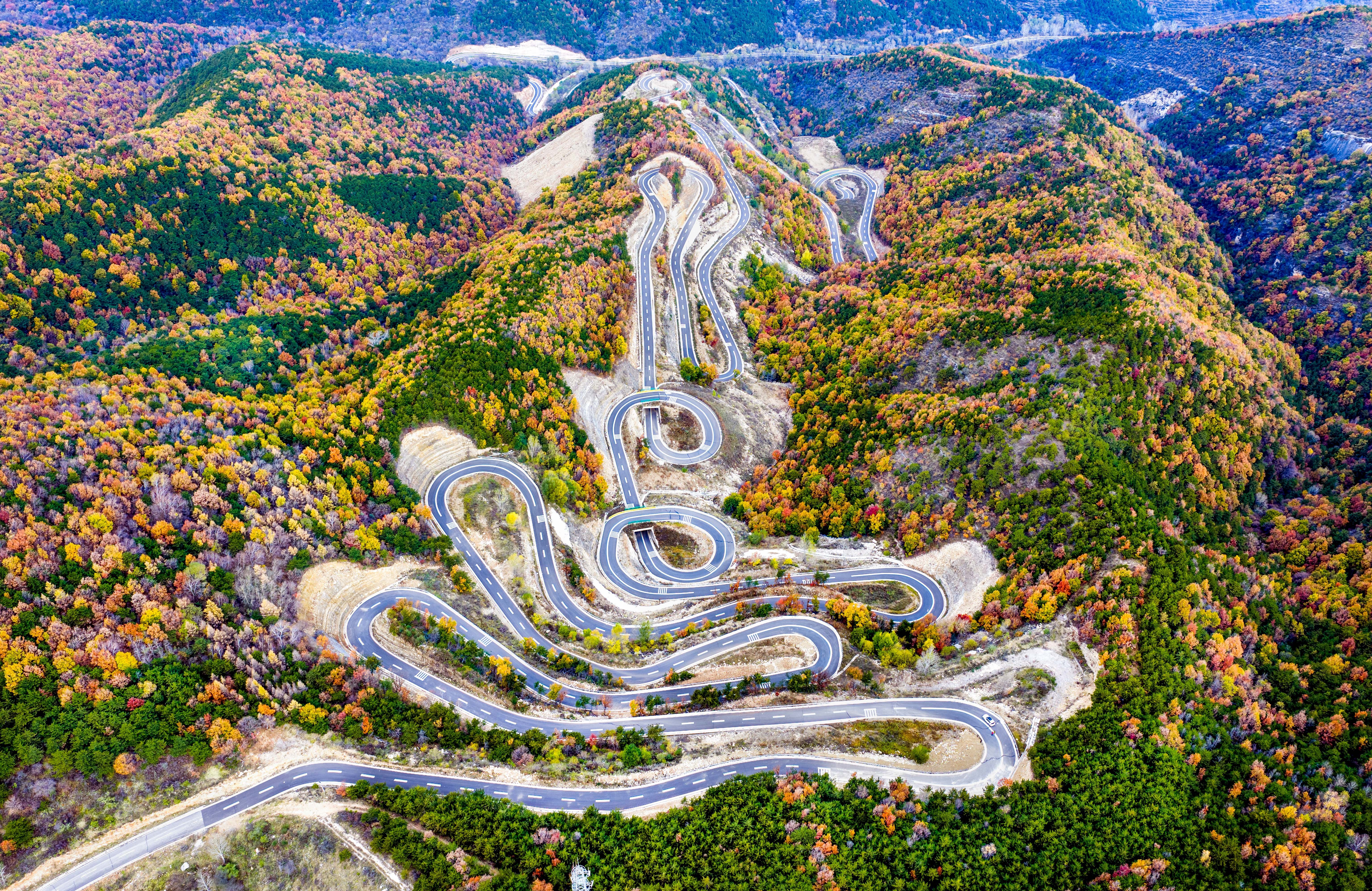 An aerial view of a road with many sharp switchbacks on a mountainside covered in autumn-colored trees