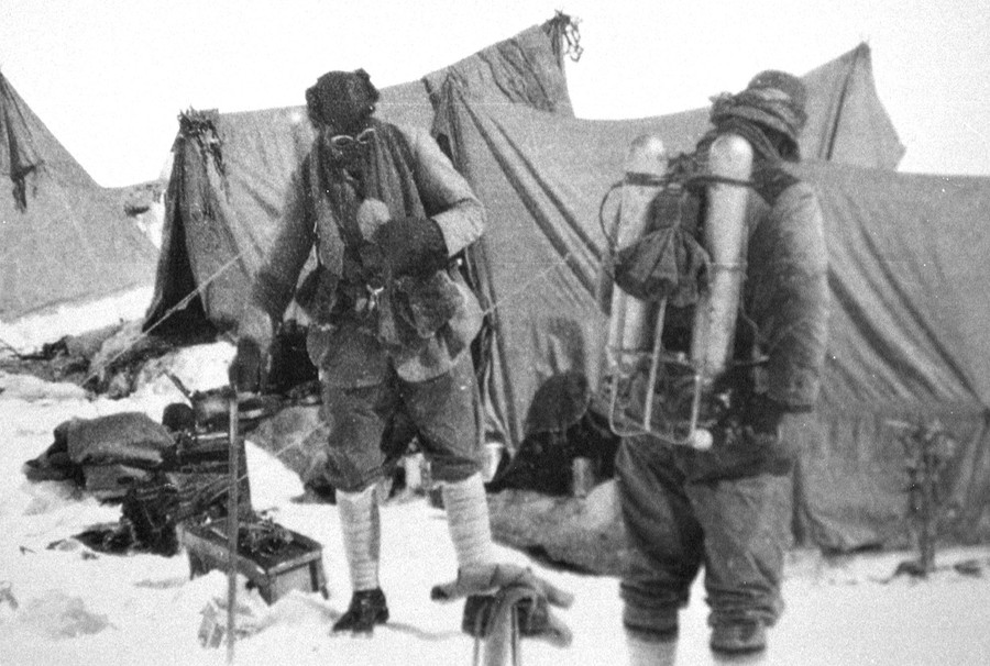 Two mountain climbers wear oxygen tanks near tents at a base camp.