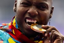 An athlete playfully bites a gold medal during a ceremony. Their canine teeth have been modified to look like long fangs.