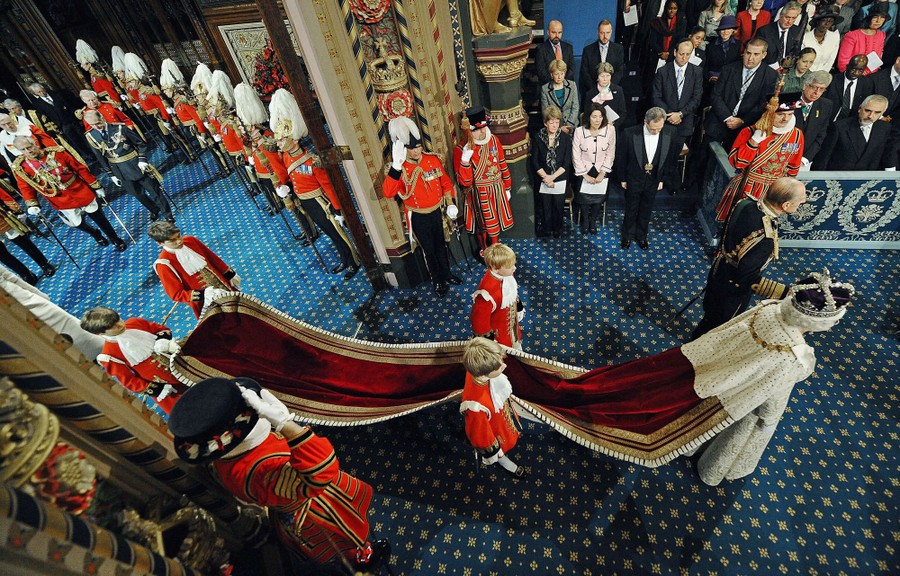 The Queen, wearing a crown and a long cape carried by four boys, walks through a crowd after giving a speech.