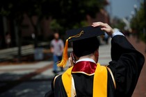 The back of a graduation in a cap and gown. He is talking on his cell phone. 