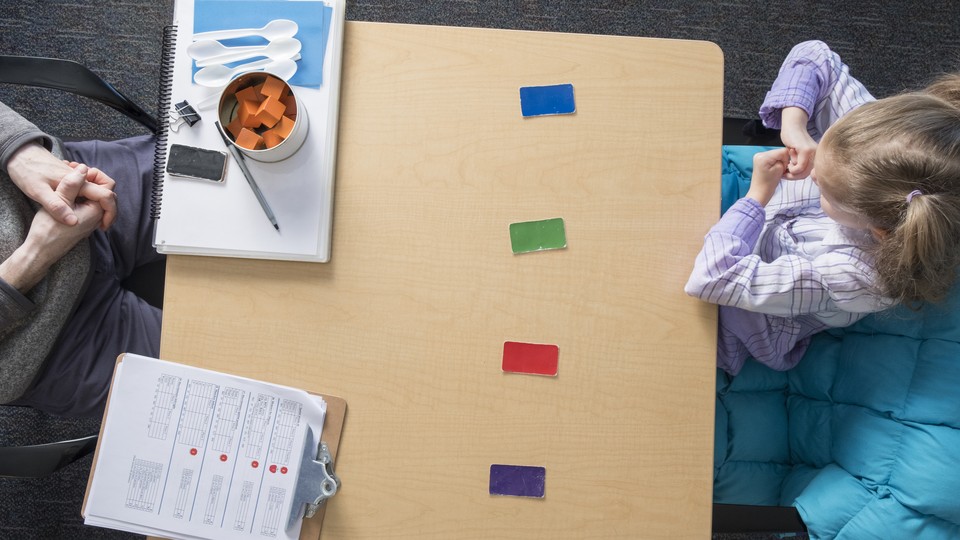 A child and an examiner sit at a table that includes colored blocks, a clipboard, and plastic spoons.