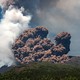 A large plume of brownish-colored ash rises from the top of a volcano.