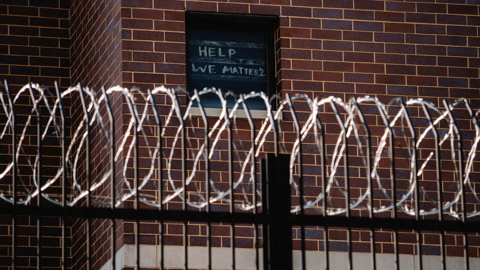 Barbed-wire in the foreground of a jail. A sign in the window reads, 'Help We Matter 2'