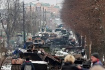 Several destroyed military vehicles sit in the middle of a road in a town.