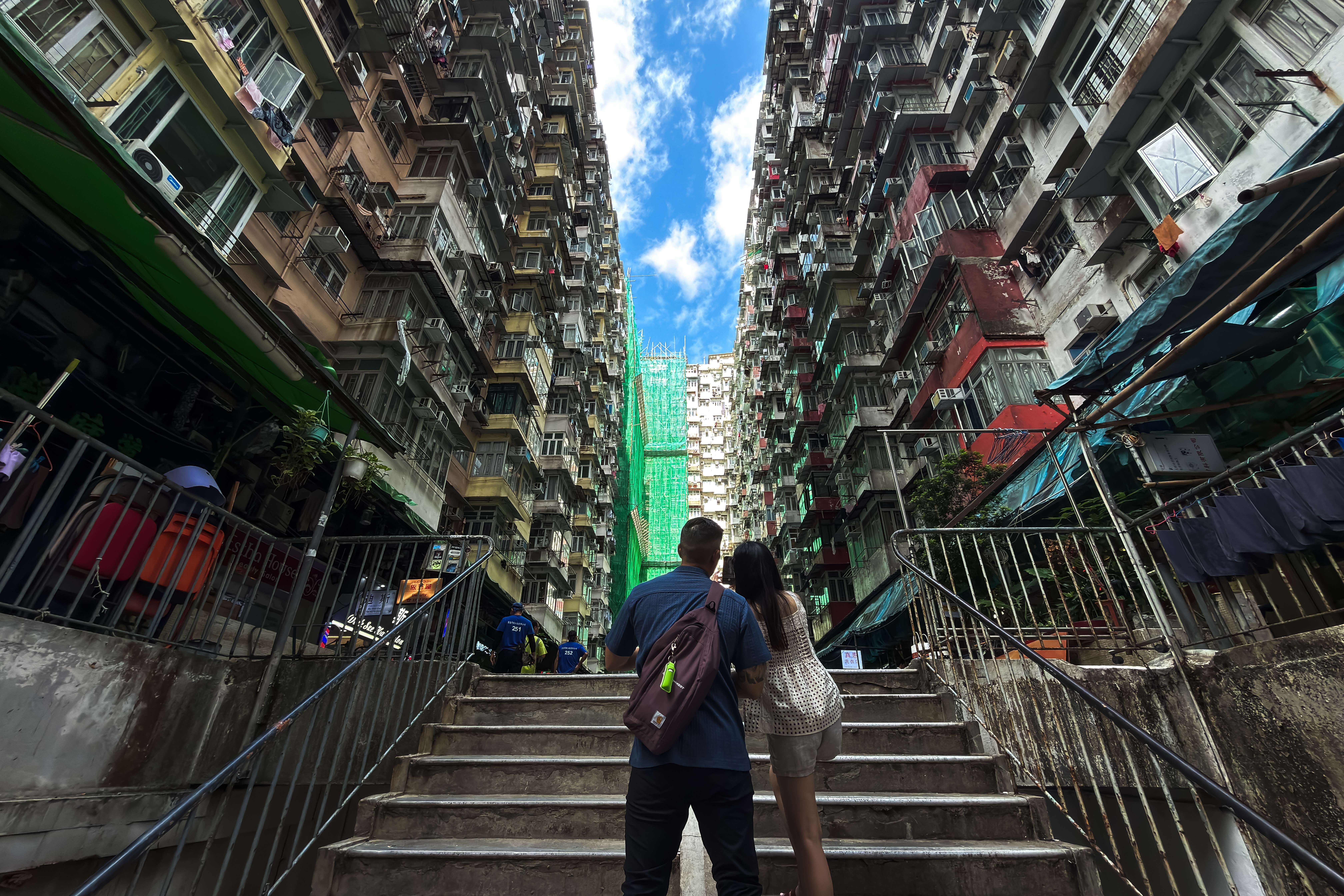 Two people stand on stairs, looking up at the sky between many tall, densely-built residential buildings.