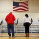 The backs of voters as they cast their ballots. They stand underneath an American flag, presumably in a gymnasium.