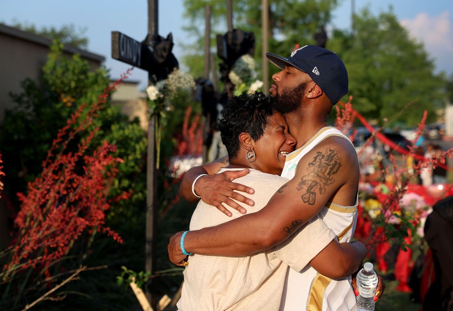 A mother and son embrace, emotional, standing in front of a makeshift memorial.
